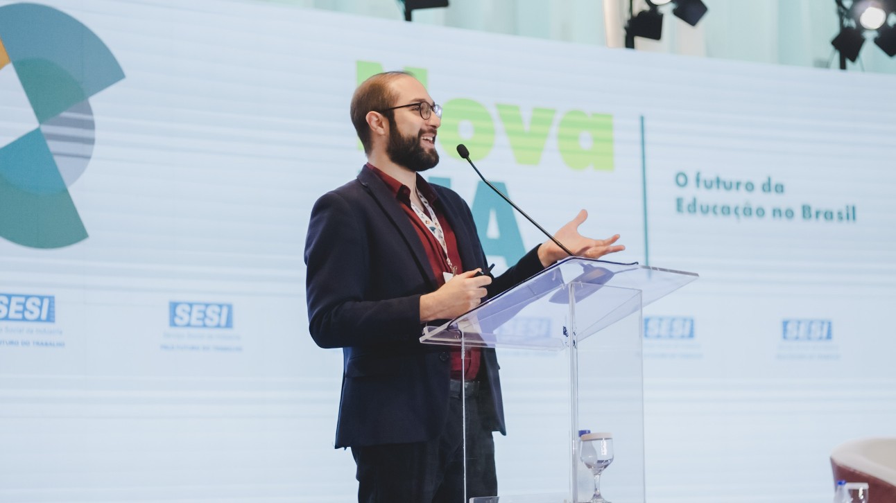Uma pessoa falando em um pódio com um banner atrás dela. A pessoa é o presidente do SESI, Eduardo Eugenio Gouvêa Vieira, e está apresentando o projeto Nova, que visa promover a inovação e a qualidade na educação no Brasil. O banner atrás dele é azul e verde com a palavra “nova” escrita nele. O banner também tem as palavras “O futuro da Educação no Brasil” escritas nele. O pódio é feito de plástico transparente e tem um copo de água em cima dele. O fundo é uma sala branca com uma grande janela.