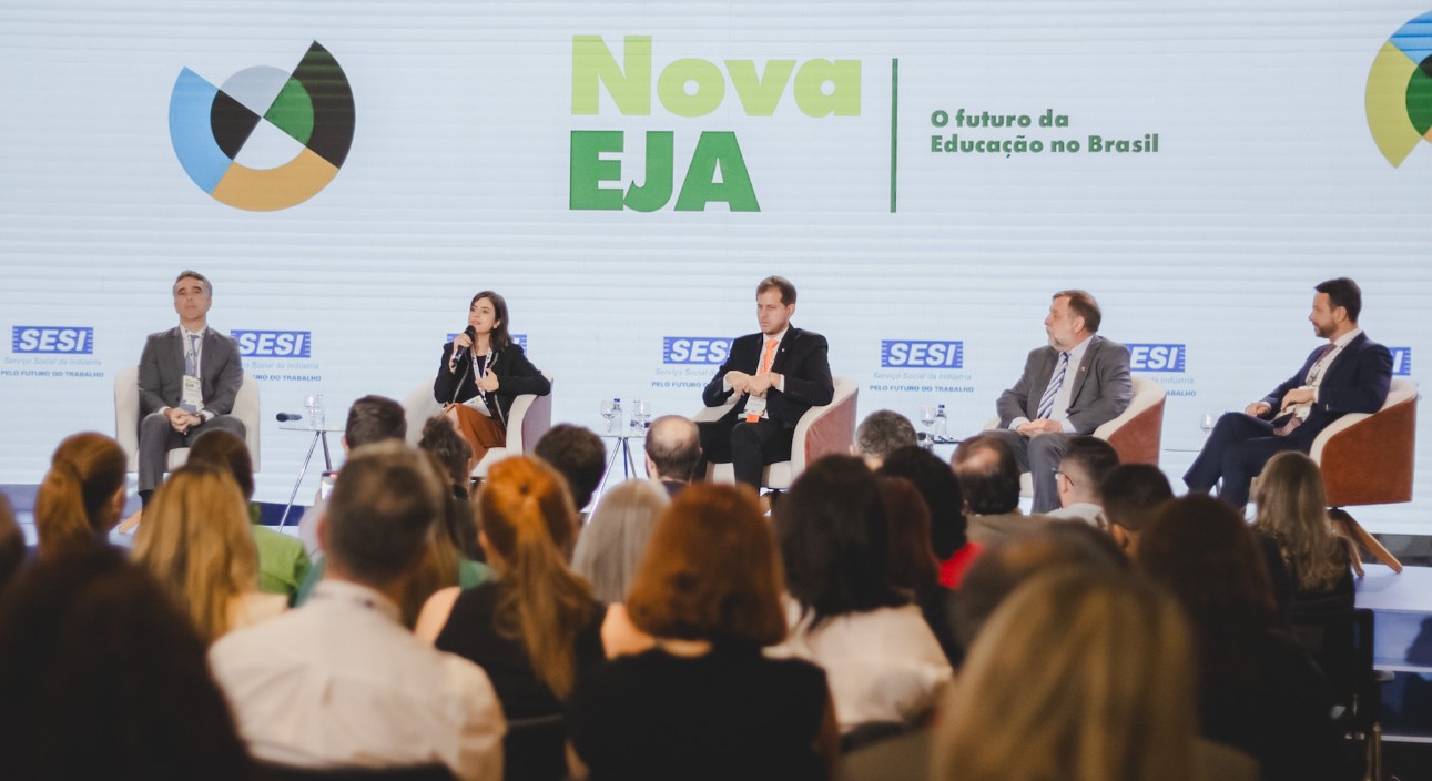 Uma foto de uma discussão em painel no evento Nova EJA. Os participantes são, da esquerda para a direita, o secretário de Educação do Estado de São Paulo, Rossieli Soares, o presidente do SESI, Eduardo Eugenio Gouvêa Vieira, a diretora do ESI, Ana Lúcia Almeida Gazzola, o presidente da Saab, Micael Johansson, e o presidente da Ericsson, Börje Ekholm. Eles estão sentados em um palco com uma grande tela atrás deles. A tela lê “Nova EJA” e “O futuro da Educação no Brasil”. A plateia é visível em primeiro plano. O fundo consiste em uma parede branca com os logos do SESI e do ESI.