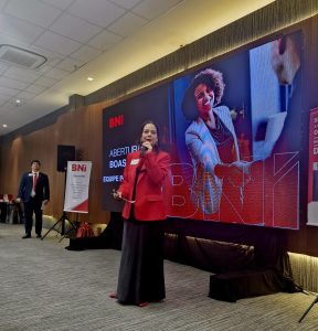 Mulher falando ao microfone durante apresenta&ccedil;&atilde;o do BNI Para&iacute;ba, com tel&atilde;o ao fundo exibindo mensagem de boas-vindas.